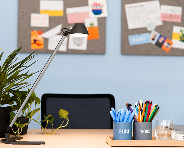 Moderner Schreibtisch mit Lampe, Pflanzen und Stiftehalter vor einer blauen Wand mit Pinnwand