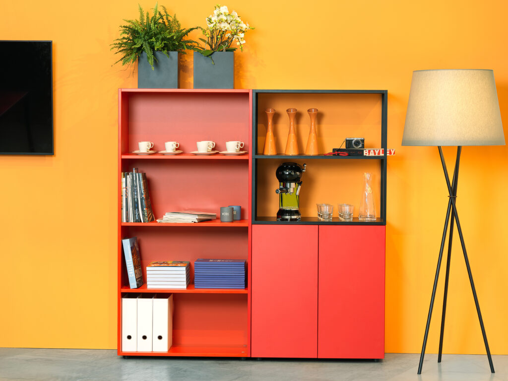 Offenes Regal mit Tassen, Büchern, Pflanzen und Kaffeemaschine vor einer warmen Wandfarbe