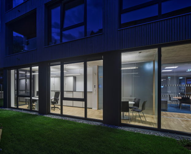 Glasfront mit Einblick in einen Bürobereich mit Besprechungsecke und Arbeitsplätzen, beleuchtet bei Nacht.
