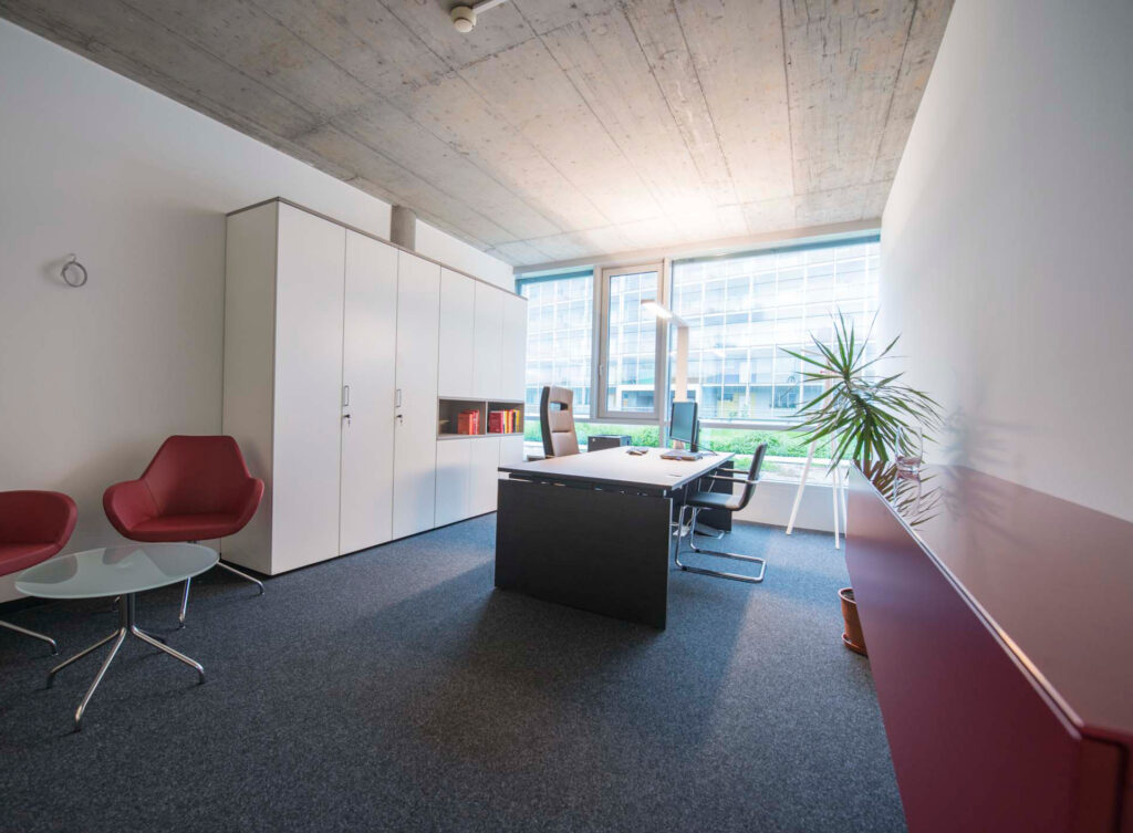 Lichtdurchflutetes Einzelbüro mit schwarzem Tisch, weißen Schränken und roten Lounge-Sesseln